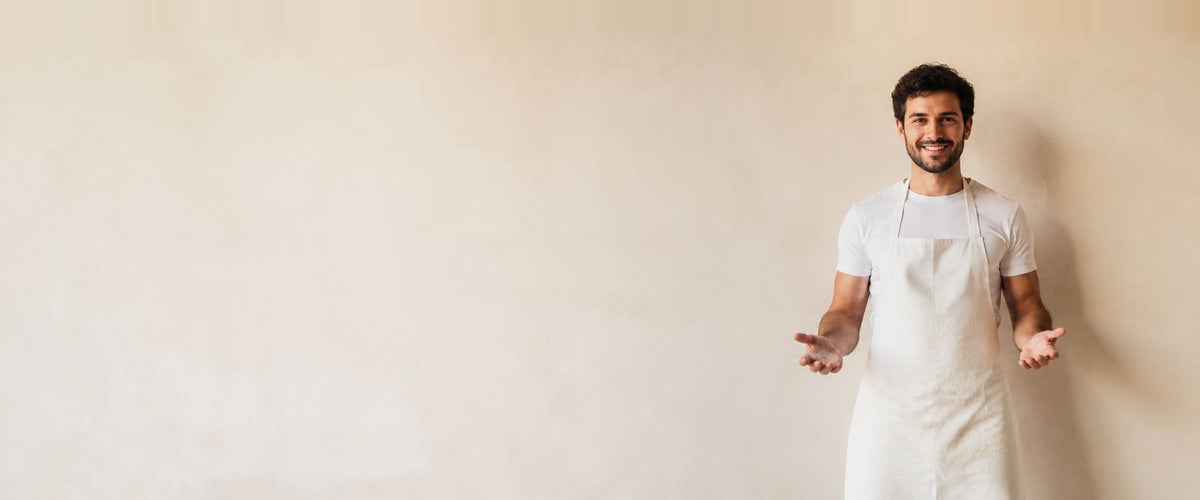 Homme en t-shirt blanc portant un long tablier à bavette écru, les bras ouverts.