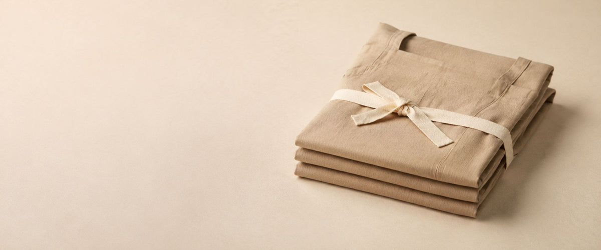 Pile de trois tabliers pliés en toile beige, entourée d'un ruban ivoire noué.