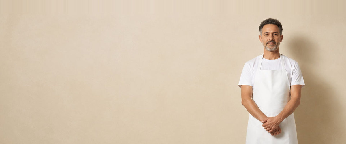 Homme en t-shirt blanc portant un long tablier à bavette blanc, les mains jointes.