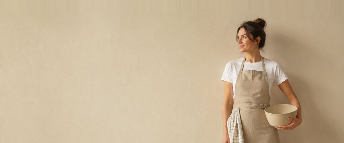 Femme en tablier à bavette beige, un torchon à carreaux à la ceinture et un saladier sous le bras.