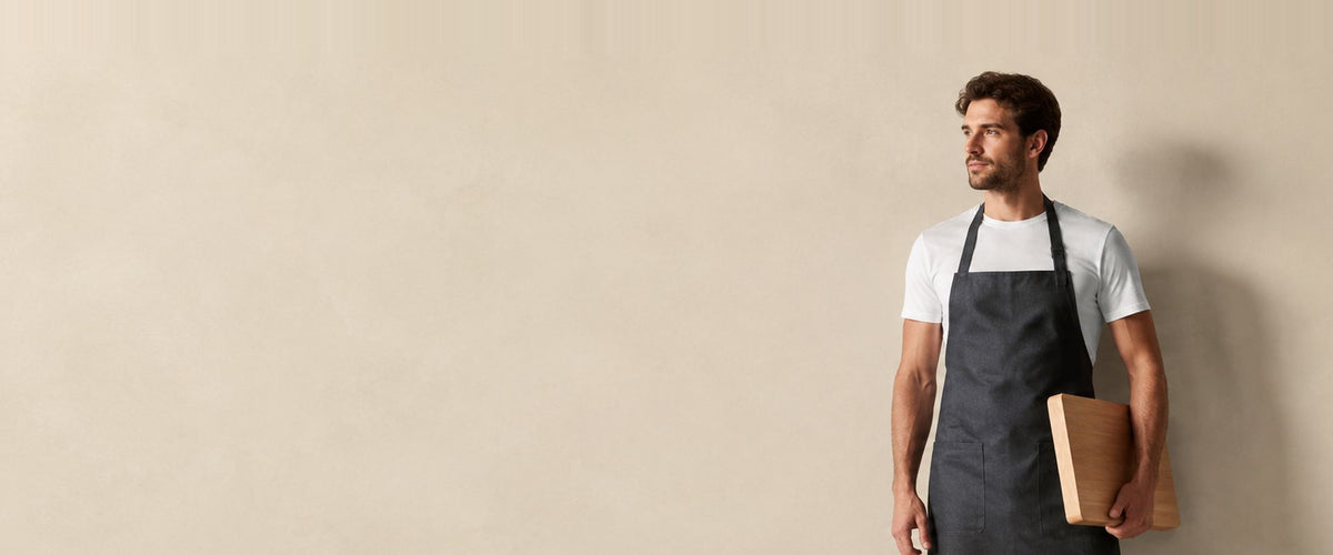 Homme en t-shirt blanc portant un tablier à bavette gris ardoise, une planche à découper sous le bras.