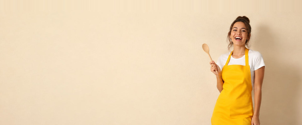 Femme riant en tablier à bavette jaune vif, une cuillère en bois à la main.