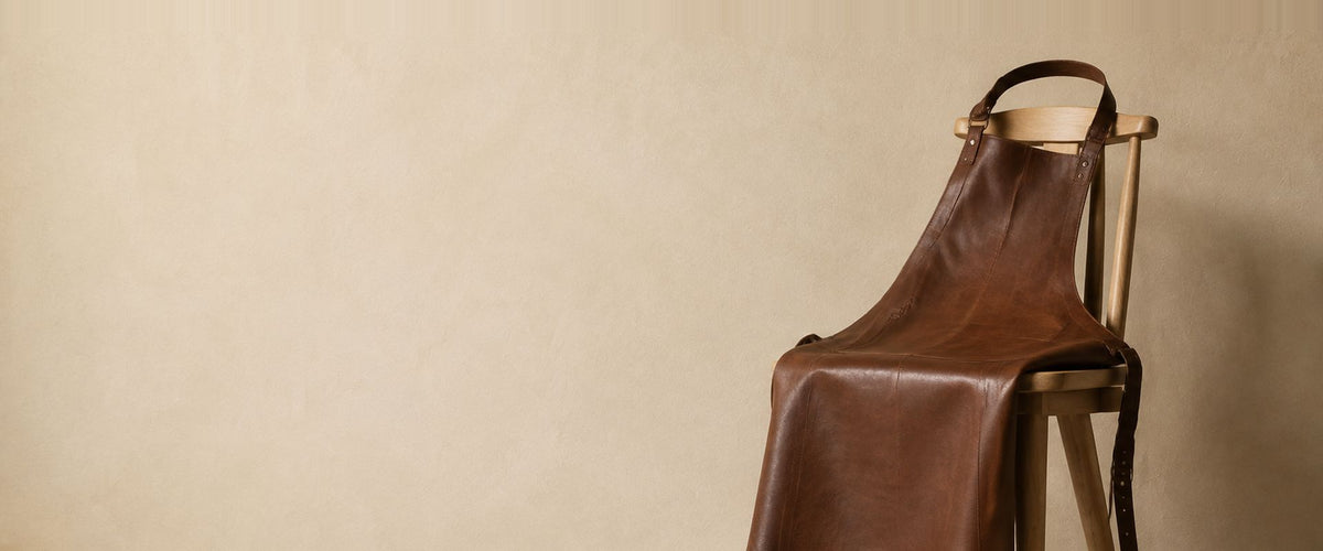 Tablier à bavette brun d'aspect cuir, posé sur le dossier d'une chaise en bois.