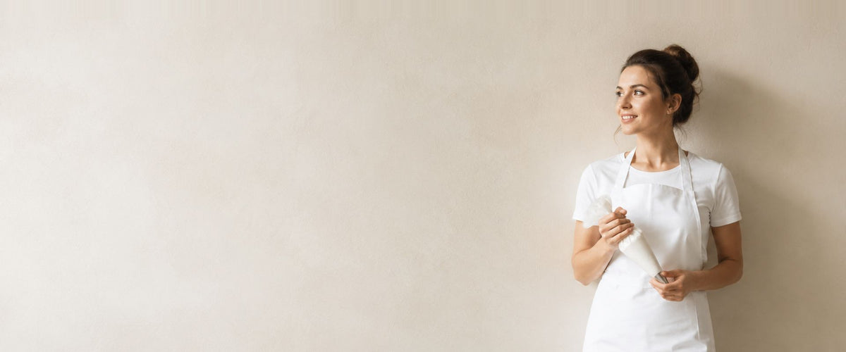Femme en veste blanche et long tablier blanc, un torchon roulé entre les mains.